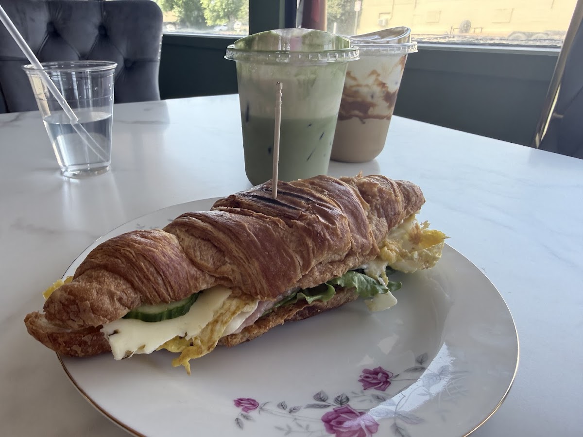 A croissant sandwich on floral china beside an iced matcha latte and a churro iced coffee